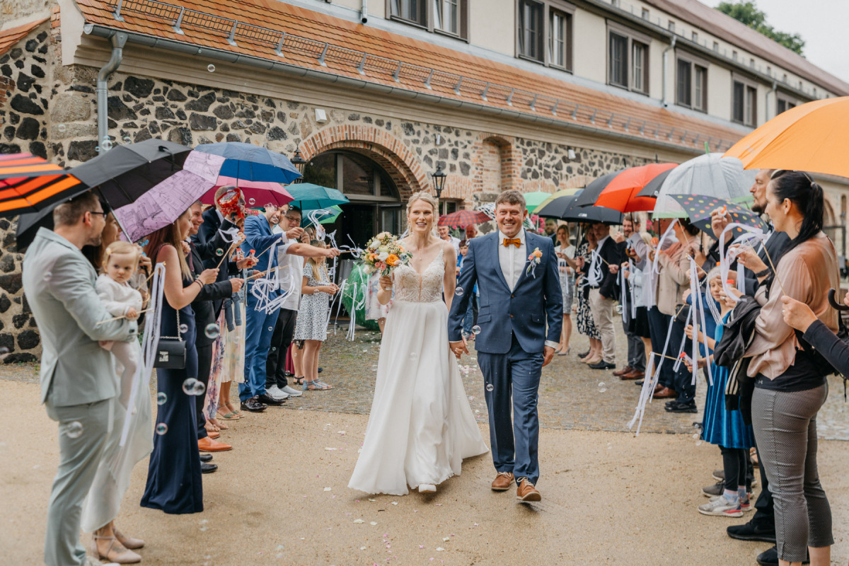Sandra und Andy verlassen das Gut am See unter bunten Regenschirmen