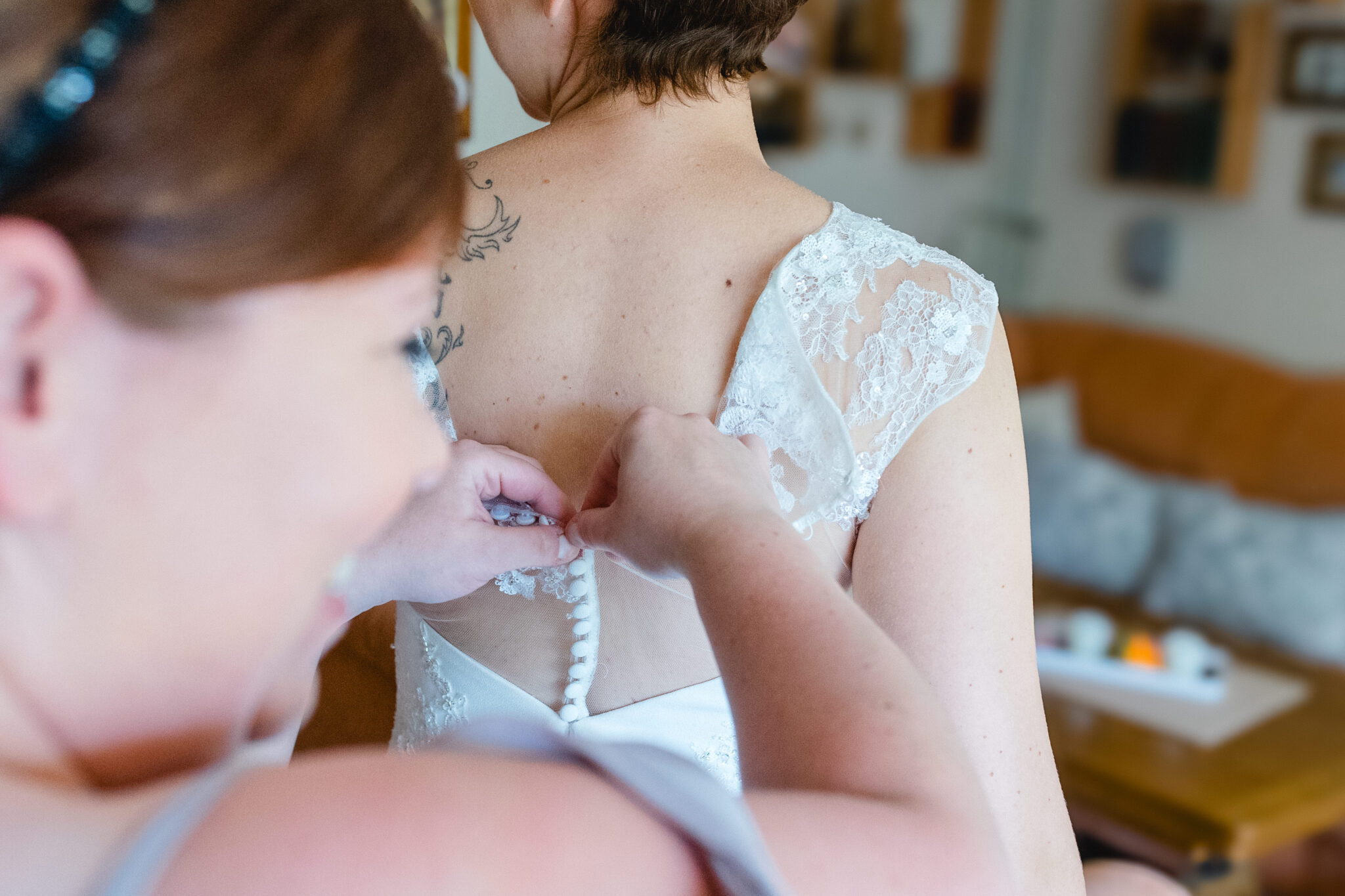 Eine Begleitperson schließt die Knopfleiste am Spitzenkleid der Braut beim Getting Ready